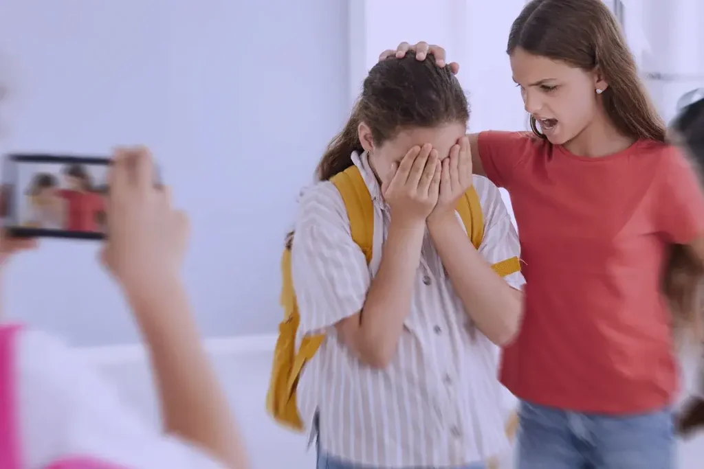 Scena di bullismo a scuola: una ragazza urla contro una compagna che si copre il viso in lacrime, mentre un'altra studentessa riprende la scena con il telefono.