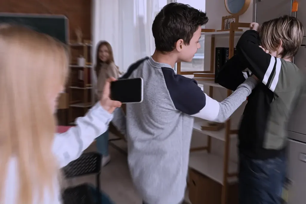 Scena di bullismo a scuola: un ragazzo spinge un compagno contro gli armadietti mentre una studentessa riprende la scena con il cellulare