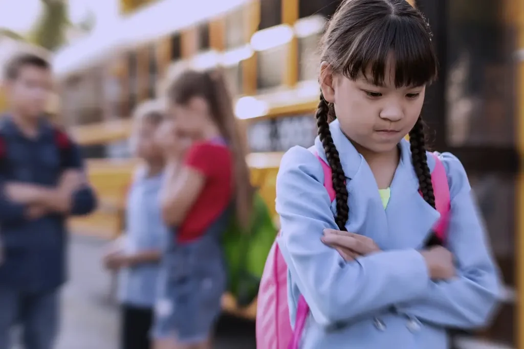 Bambina con le braccia incrociate e sguardo triste vicino a uno scuolabus, mentre un gruppo di compagni parla in lontananza.