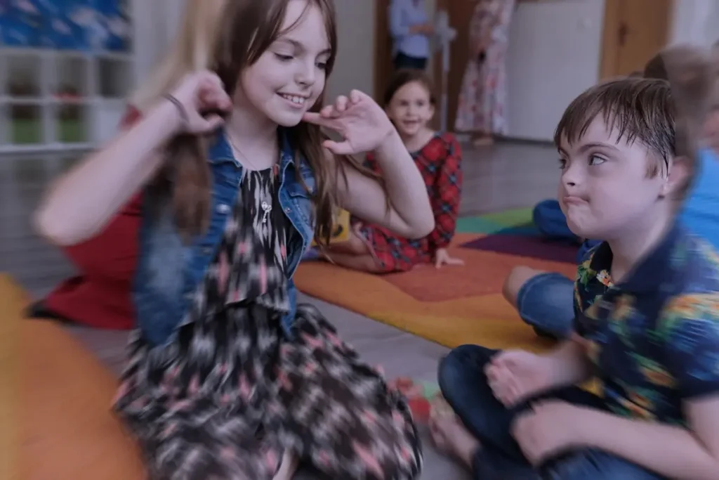 Bambina e bambino con disabilità intellettiva seduti a terra in un'aula scolastica, che interagiscono tra loro in un momento di gioco o attività educativa.